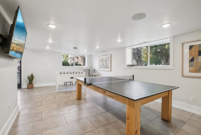 Modern recreational room with ping pong and foosball tables, bright windows, and a large wall-mounted TV for entertainment.