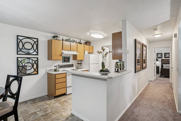 Dog-Friendly Apartments in Ogden, UT - Pinebrook Apartments - Kitchen With Light Wood Cabinets, White Appliances, Breakfast Bar, Tile Flooring, And Hallway Leading To Bedroom.