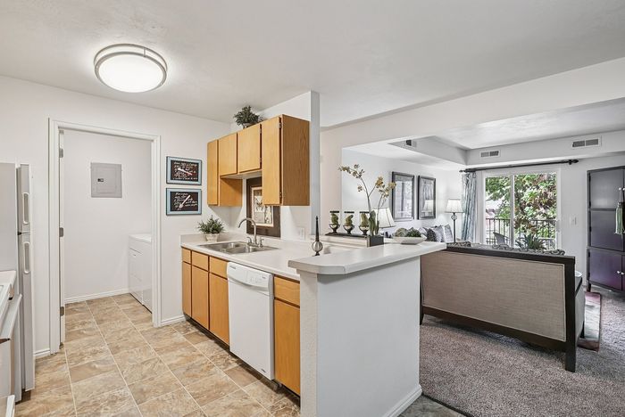Bright apartment kitchen with modern white countertops, wooden cabinets, and seamless flow into a cozy living area with elegant decor and large windows.