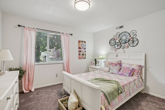 Ogden, UT, Pet-Friendly Apartments - Pinebrook Apartments - Bedroom With White Bed Frame, Patterned Bedding, Large Window With Pink Curtains, Carpet, And Decorative Wall Art.