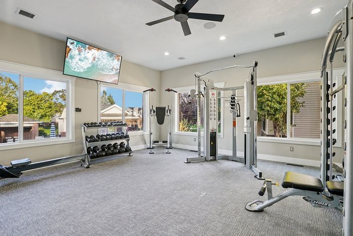 Bright, modern gym in an apartment complex with large windows, giving a vibrant outdoor view. Equipped with diverse fitness machines and a sleek dumbbell rack.