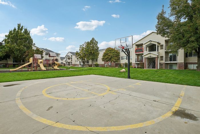 Vibrant apartment community featuring modern buildings, a playground for children, and a basketball court, set against a backdrop of lush green lawns and clear blue skies.