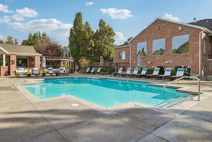 Stunning community pool area surrounded by inviting sun loungers, lush greenery, and charming brick architecture under a bright blue sky.