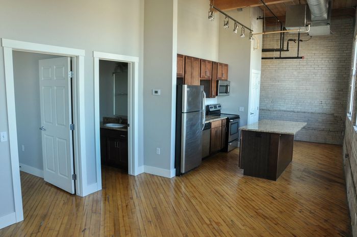 Modern apartment kitchen with stainless steel appliances, sleek wooden cabinets, and exposed brick and beam accents.