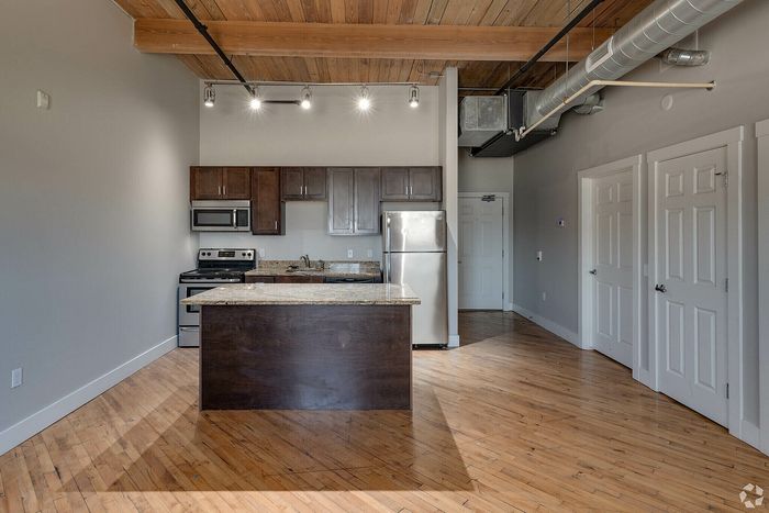 Modern loft-style kitchen with sleek stainless steel appliances, warm wood cabinets, and polished hardwood floors under industrial-chic exposed ductwork.