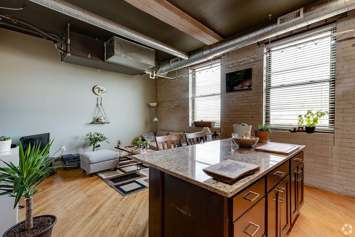 Charming industrial-style apartment with open-concept living and kitchen area, featuring exposed brick walls, large windows, and modern decor.