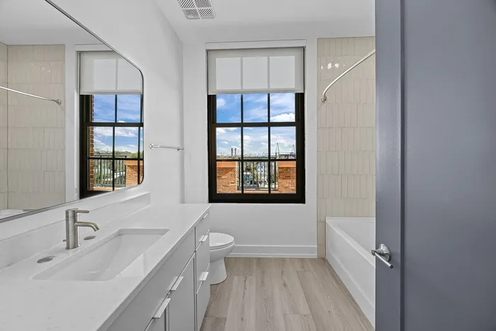 Bright, modern bathroom with a large window offering a scenic city view, sleek vanity with white countertop, and an inviting bathtub.