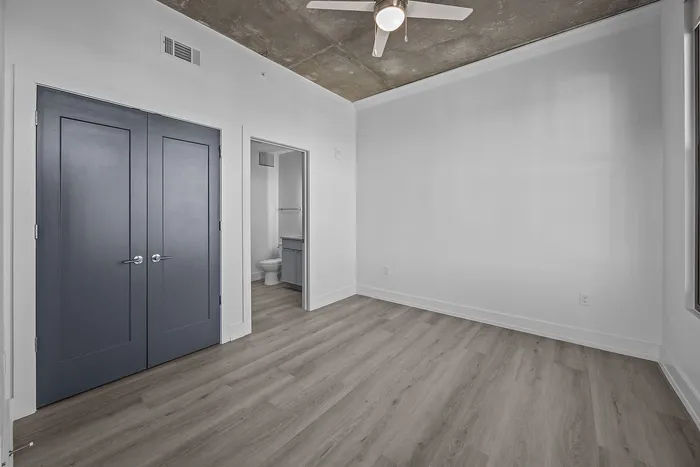 Bright modern room with sleek wood flooring, featuring a stylish ceiling fan and dark double doors leading to a private bathroom.