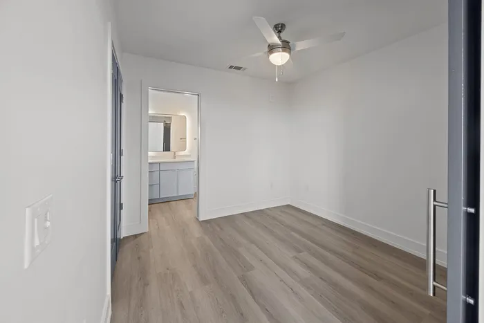 Inviting room with modern ceiling fan, sleek wood flooring, and a glimpse into a stylishly lit bathroom.
