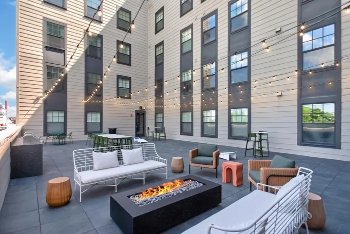 Chic urban patio featuring cozy seating around a modern fire pit, framed by elegant string lighting and a stylish apartment backdrop.