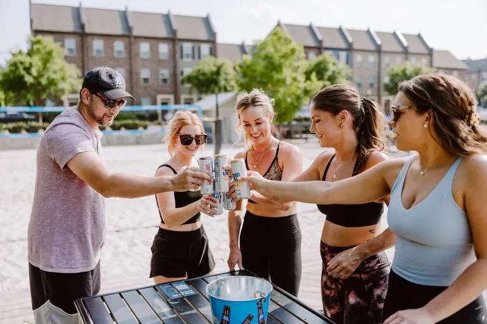 Group of friends enjoying a sunny outdoor gathering, toasting with drinks by a sand volleyball court, showcasing a vibrant community lifestyle.