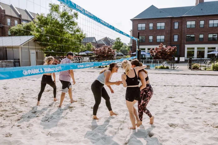 Enthusiastic group enjoying a sunny day playing beach volleyball on a sandy court, surrounded by lush greenery and modern brick buildings.