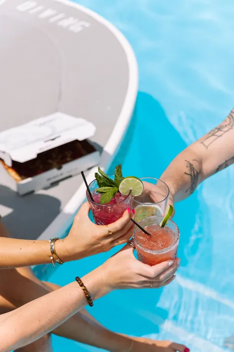 Vibrant poolside scene with friends toasting refreshing cocktails next to a delicious pizza box, creating the perfect summer vibe.