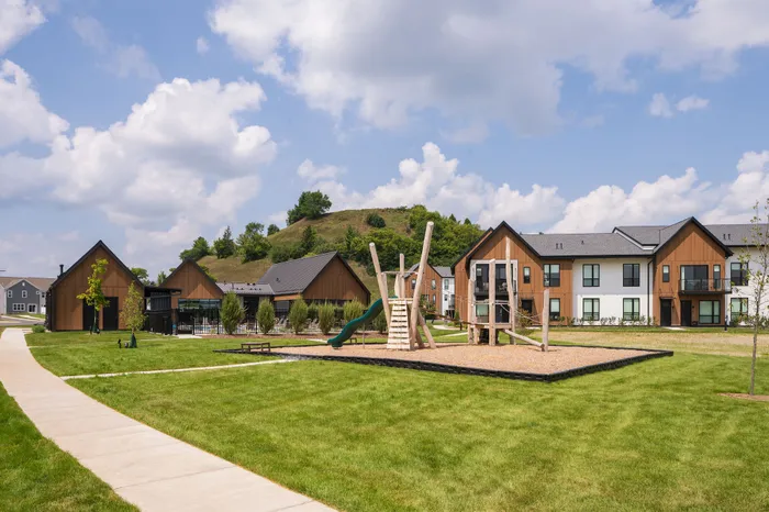 Charming modern apartment complex with rustic design, featuring lush green lawns and a fun playground, set against a scenic hillside backdrop.
