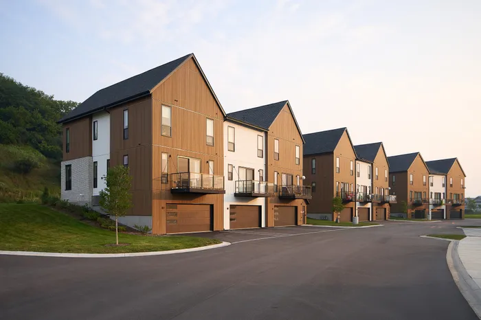 Charming row of modern townhomes with stylish wooden facades, featuring private balconies and two-car garages, set against a serene natural landscape.