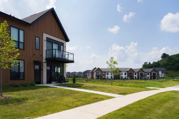 Modern apartment complex surrounded by lush green lawns and a scenic hillside, featuring sleek architectural design under a vibrant blue sky.