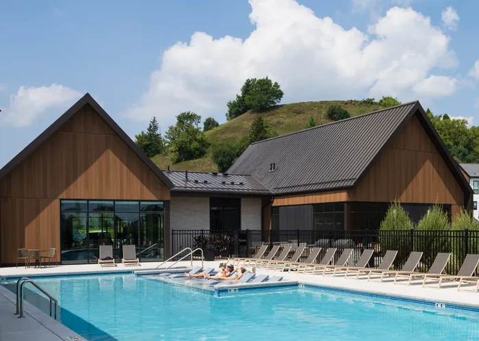 Inviting outdoor pool area lined with sun loungers, adjacent to modern wooden-clad buildings set against a lush hillside backdrop.