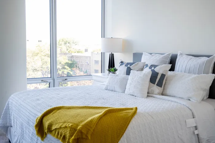 Bright and airy bedroom showcasing a luxurious bed with crisp white linens and vibrant yellow accents, framed by large windows with a stunning view.