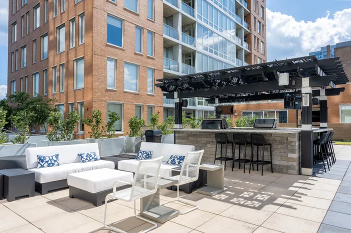 Chic outdoor lounge area with modern white seating, decorative pillows, and a stylish bar under a pergola, set against a contemporary brick building.