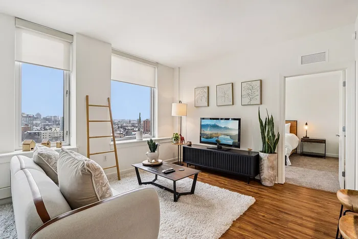 Bright and modern living room with large windows offering city views, featuring a cozy setup with a plush couch, stylish decor, and a sleek TV unit, seamlessly connecting to a comfortable bedroom.