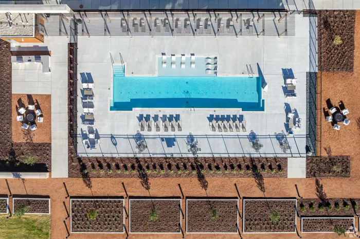 Aerial view of a modern apartment complex with a sleek swimming pool surrounded by stylish lounge chairs, lush landscaping, and outdoor seating areas.