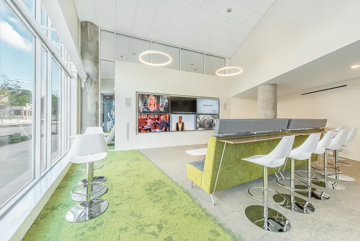 Modern and inviting lounge area with sleek bar stools, a green sofa with high backrests, and a wall-mounted multi-screen display. Large windows offer ample natural light, complemented by stylish circular overhead lighting.