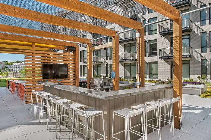 Modern outdoor lounge area with sleek bar seating and TV, framed by stylish wooden pergola, offering a perfect entertainment spot in a contemporary apartment complex.