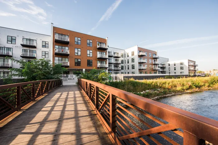 Modern riverside apartments with spacious balconies, surrounded by lush greenery, connected by a charming wooden bridge, offering serene water views.