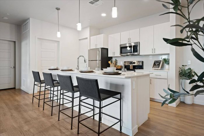 2-Bedroom Apartments in East Mesa, AZ - Acero Eastmark - Modern White Kitchen Featuring Island Seating, Pendant Lights, Stainless Steel Appliances, and Wood-Style Flooring.