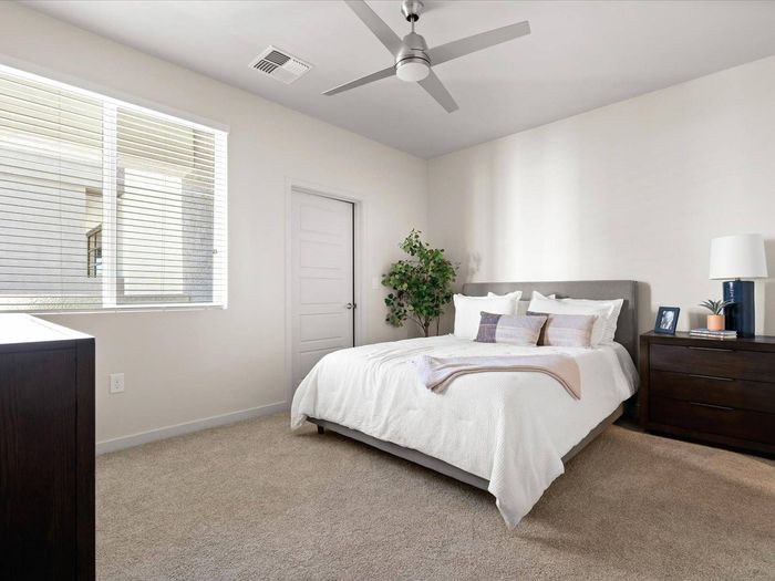 3 BR Apartments in Glendale AZ - Acero at the Stadium - Carpeted Bedroom with a Large Bed, White WAlls, Wooden Dresser and Nightstand Cabinet, Ceiling Fan, and a Window