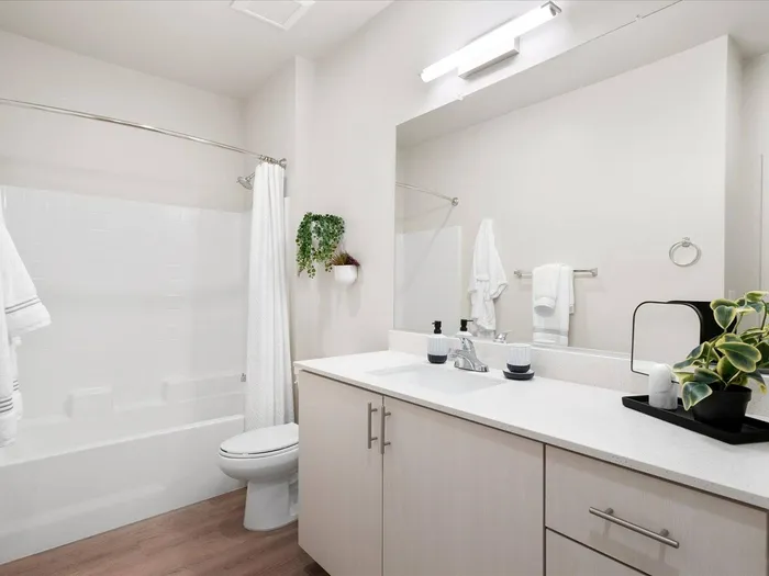 Glendale, AZ, Apartments - Acero at the Stadium - Bathroom with White Quartz Counter Sink, Off-White Cabinetry, a Large Mirror, and a Shower with a Tub and White Shower Curtain.