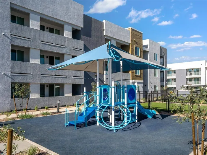 2-Bedroom Apartments in Glendale, AZ - Acero at the Stadium - Outdoor Playground Area in Front of Property Building with a Blue and White Slide Set with an Umbrella Top on Dark Soft Rubber Turf Next to Fenced in Grass Area.