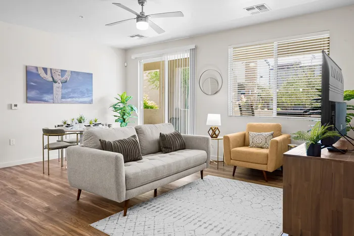 Pet-Friendly Apartments in North Phoenix AZ - Acero North Valley - Furnished Living Room with grey Couch, Yellow-Tan Armchair, White Carpet, Wood Floors, Framed Mirror and Art, Wooden TV Stand with TV, Several House Plants, Dining Table with Chairs and Place Settings, Large Windows with Blinds, and Patio Access