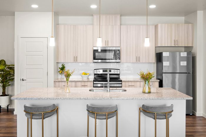 North Phoenix AZ Apartments for Rent - Acero North Valley - Modern Kitchen with Stainless Steel Appliances, Granite Countertops, Cooking Island with Hanging Lights and Stool Seating, White Tile Backsplash, Light Wood-Style Cabinetry, Pantry Door, Dark Wood Flooring, and View of a Nearby House Plant