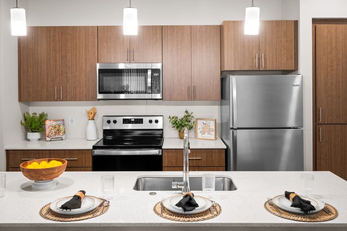 Queen Creek, AZ Apartments - Acero Harvest Station - View Of The Beautiful Kitchen From Behind The Kitchen Island Bar Area With Three Plates Set, The Kitchen Has Stainless Steel Appliances And Modern Style Wood Cabinetry