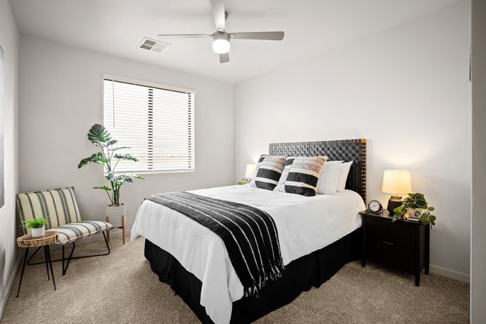 Eastmark, AZ, Apartments Near Mesa Gateway - Acero Hawes Crossing - Modern Bedroom With Ceiling Fan, Window Blinds, Upholstered Headboard, And Nightstand Lighting.