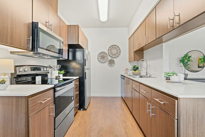 Apartments For Rent In Ridgefield, WA - Acero Ridgefield - Kitchen With Stainless Steel Appliances, Brown Hardwood Cabinets, Quartz Counters, And Vinyl Plank Flooring