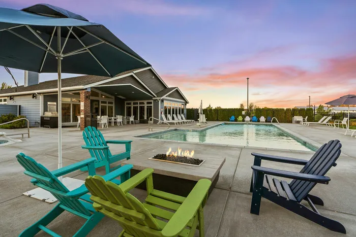 Apartments for Rent in Ridgefield, WA - Acero Ridgefield - Fire Pit Lounge With Adirondack Chairs Around A Gas Flame Table, Pool Deck Umbrellas, and Clubhouse Patio in the Background.