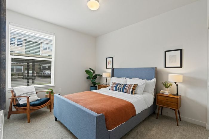 1 BR Apartments in Vancouver WA - Acero Jensen Park - Carpeted Bedrooom with a Bed, Wooden Armchair with Cushions, Wooden Nightstands with Drawers and Lamps, White Walls, and a Large Window
