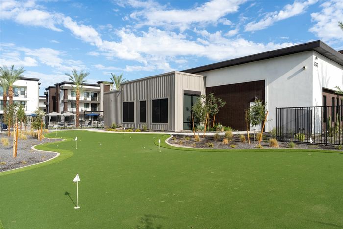 Three-Bedroom Luxury Apartments in Eastmark, AZ - Acero Hawes Crossing - Exterior Clubhouse With Putting Green, Flag Markers, Desert Landscaping, And Modern Apartment Buildings Under Blue Sky