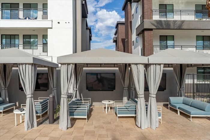2-Bedroom Luxury Apartments in Eastmark, AZ - Acero Hawes Crossing - Poolside Cabanas With Draped Curtains, Outdoor Lounge Seating, And Mounted TVs Framed By Modern Apartment Buildings.