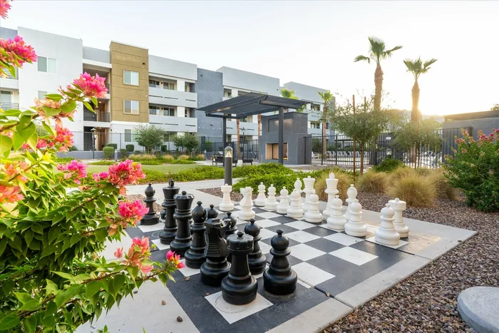 Pet-Friendly Apartments in Glendale, AZ - Acero at the Stadium - Outdoor Courtyard With Giant Chess Board, Pergola Seating, Desert Landscaping, and Apartment Buildings In The Background.