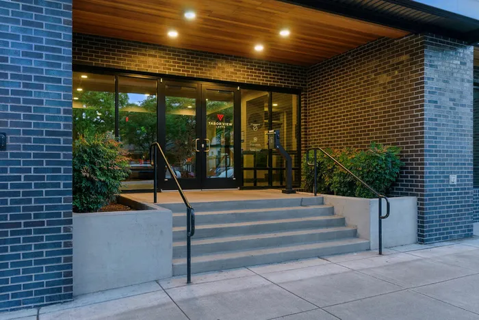 Inviting apartment entrance with modern glass doors and sleek brick exterior, surrounded by greenery.