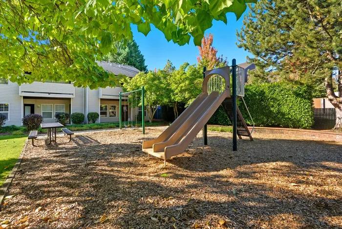 Charming community playground with a slide, surrounded by lush greenery, near cozy apartment buildings.