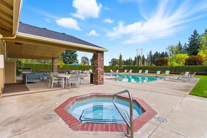 Luxurious outdoor pool area with spa and covered seating, surrounded by lush greenery and clear blue skies.