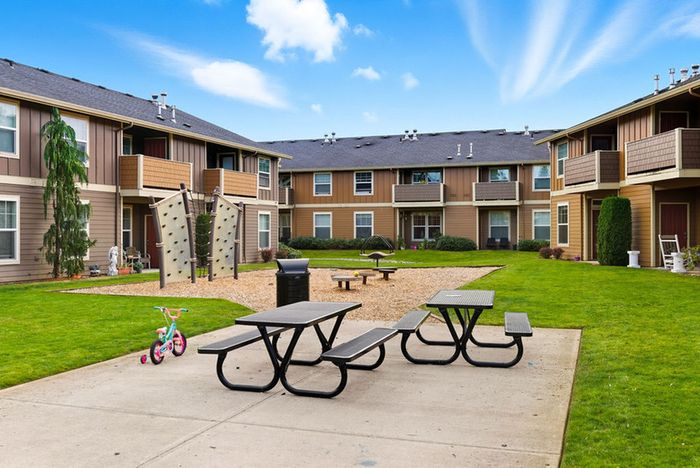 Charming apartment courtyard featuring well-maintained gardens, a children's play area, and inviting picnic tables under a vibrant blue sky.