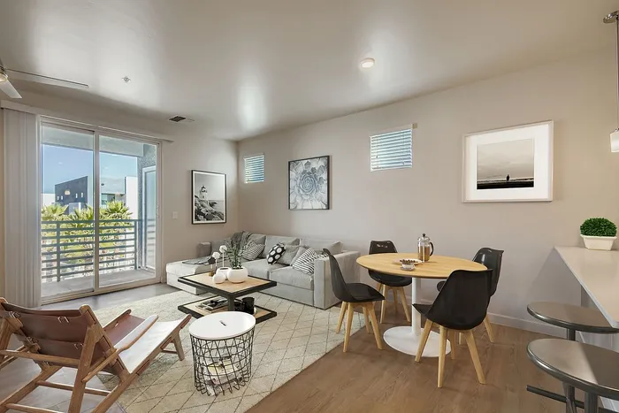 Modern living room with stylish decor, cozy gray sofa, minimalist artwork, and a round dining table, opening to a sunlit balcony with a view.