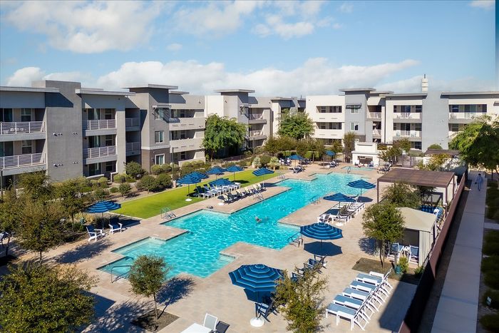 Inviting apartment complex with modern architecture features a luxurious swimming pool surrounded by sun loungers and vibrant blue umbrellas, perfect for a relaxing lifestyle.