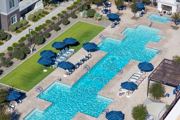 Luxurious apartment complex showcasing a sparkling swimming pool with elegant blue umbrellas and cozy lounge chairs, surrounded by well-maintained greenery.