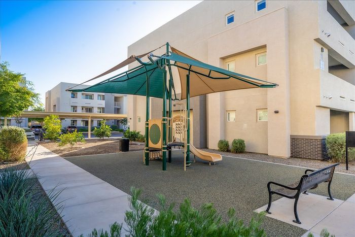 Modern apartment complex with a shaded playground area, lush greenery, and ample parking nearby.